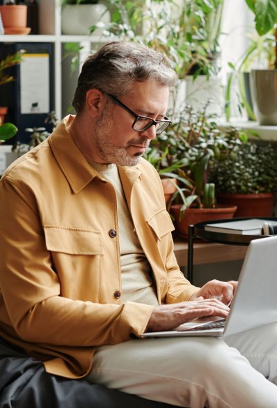 Serious mature businessman in casualwear and eyeglasses with laptop on knees networking in office while sitting on soft bag chair