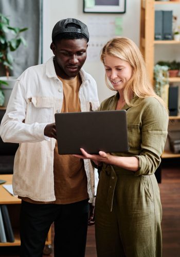 Two young intercultural employees in casualwear looking at screen of laptop held by blond businesswoman during discussion